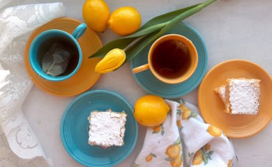 Lemon Love Note lemon cookie bars on blue and yellow tea saucers with two cups of tea in mismatched blue and yellow cups and saucers surrounded by lemons, yellow tulips, lace, and lemon dishtowels. Sugar with Spice Blog.