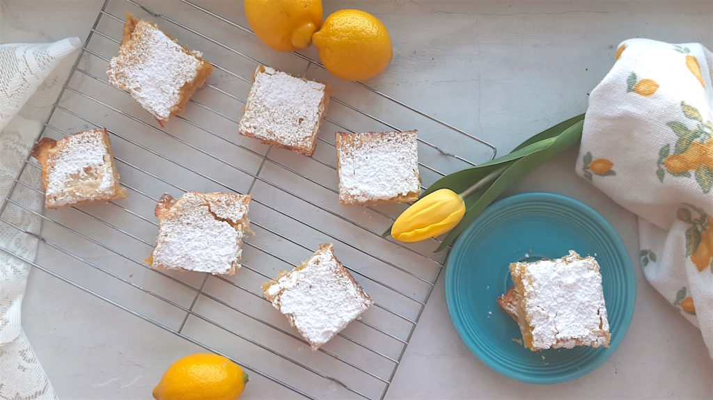 Lemon love note lemon bars cooling on a wire rack on a white background. Two love notes on a blue tea saucer surrounded by lemons, yellow tulips, and a lemon dish towel. Sugar with Spice Blog.