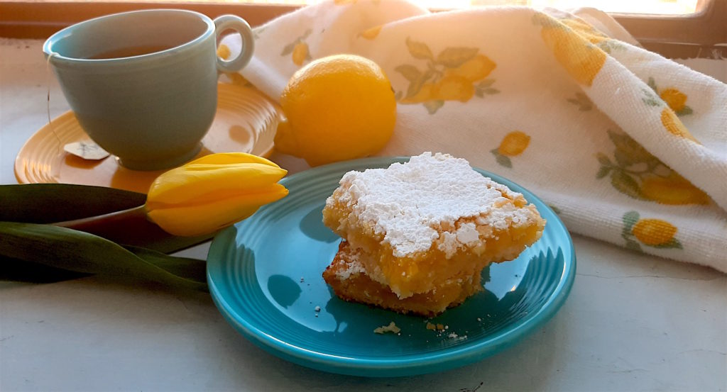 Two Lemon Love Note lemon bars on a blue saucer with a cup of tea in mismatched yellow and blue cup and saucer, with yellow tulip, lemon, and lemon dish towel on a white window sill.
