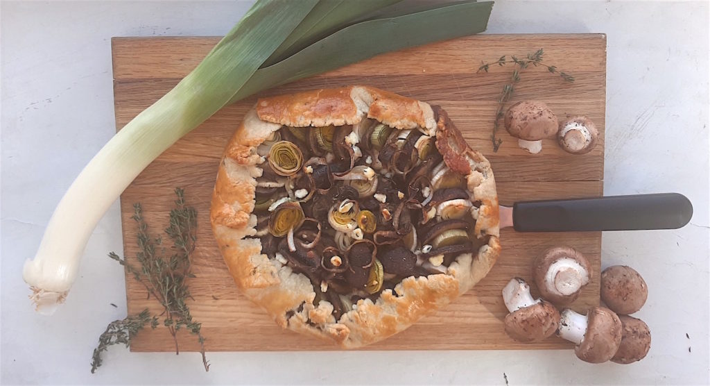 Overhead shot of Mushroom Leek Galette with goat cheese and thyme wrapped in all butter crust on a wooden cutting board. Surrounded by leeks, thyme and mushrooms. One slice cut out.