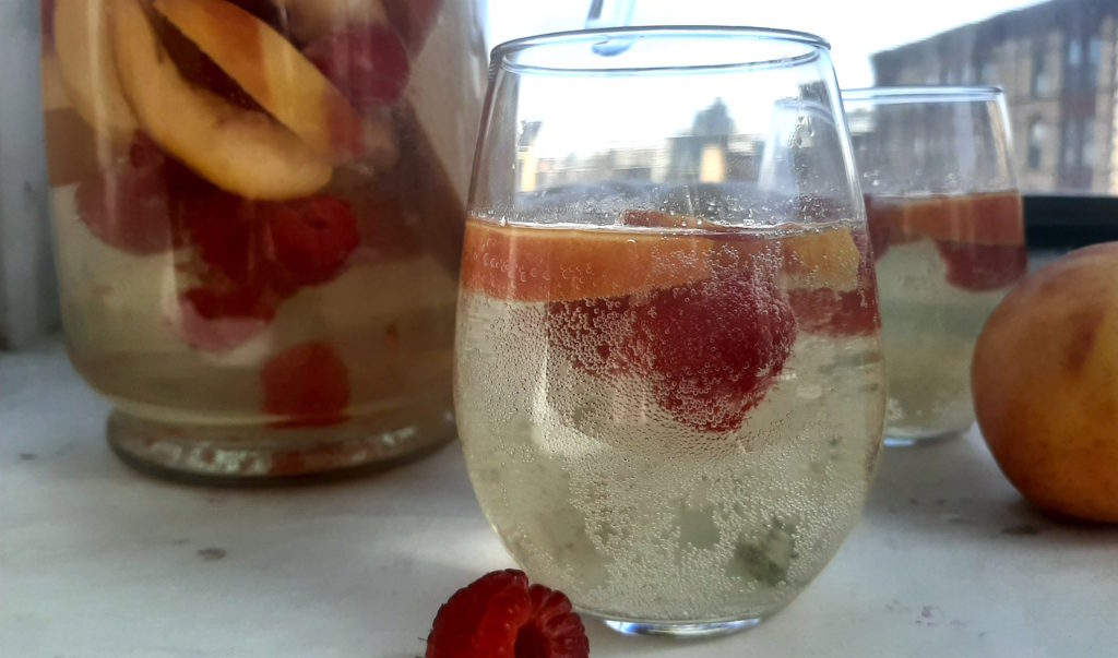 Close up of Peach Sangria in stemless wine glass with peaches and raspberries. Surrounded by fresh peaches and raspberries, full pitcher of peach sangria and a second glass in the background on a white window sill.