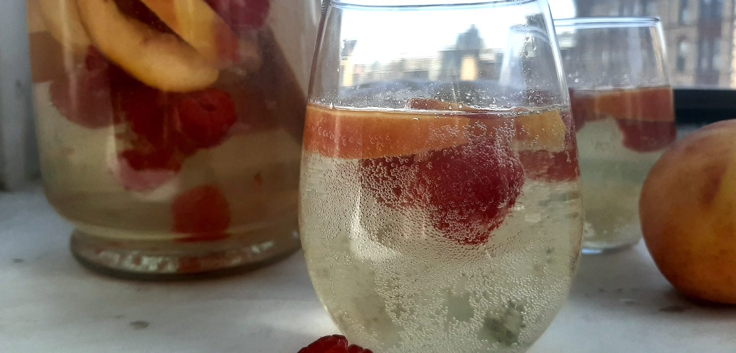 Close up of Peach Sangria in stemless wine glass with peaches and raspberries. Surrounded by fresh peaches and raspberries, full pitcher of peach sangria and a second glass in the background on a white window sill.