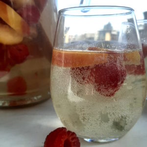 Close up of Peach Sangria in stemless wine glass with peaches and raspberries. Surrounded by fresh peaches and raspberries, full pitcher of peach sangria and a second glass in the background on a white window sill.