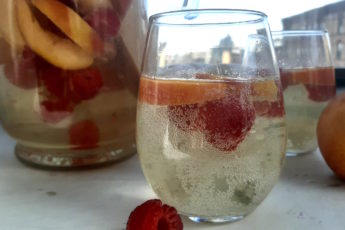 Close up of Peach Sangria in stemless wine glass with peaches and raspberries. Surrounded by fresh peaches and raspberries, full pitcher of peach sangria and a second glass in the background on a white window sill.