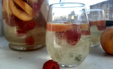 Close up of Peach Sangria in stemless wine glass with peaches and raspberries. Surrounded by fresh peaches and raspberries, full pitcher of peach sangria and a second glass in the background on a white window sill.