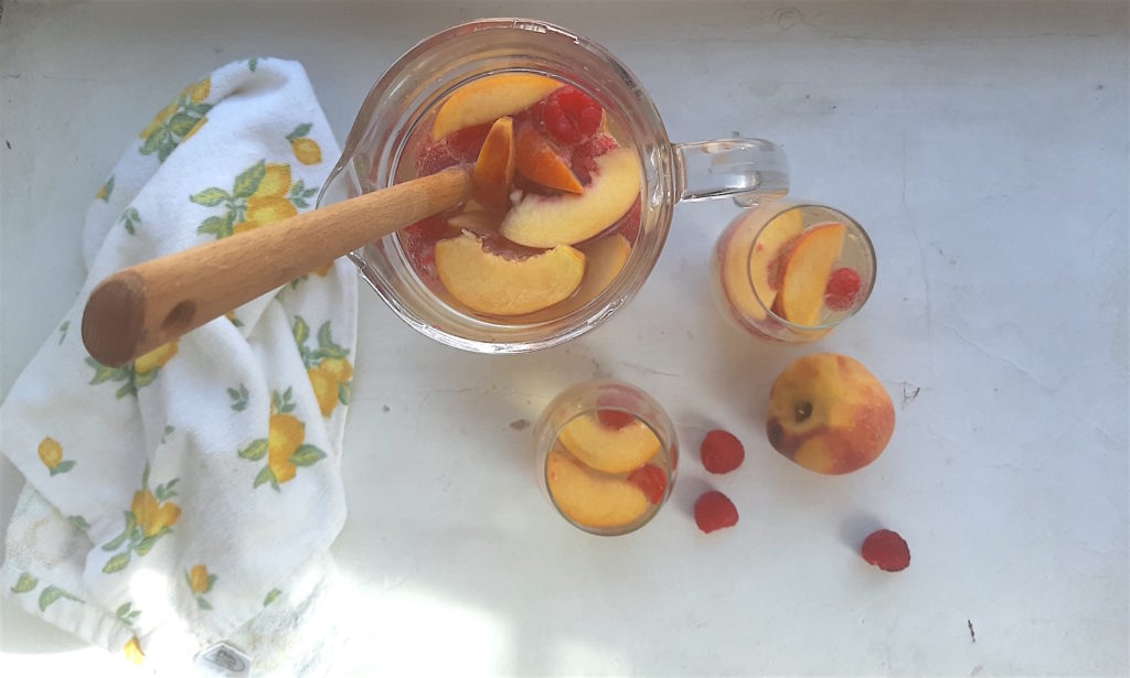 Peach sangria in a glad pitcher with fresh peaches and raspberries and a wooden spoon, next two more peach sangria and fruit in two glasses surrounded by fresh peaches and raspberries and a lemon dish towel on a white background. Overhead shot. Sugar with Spice Blog.