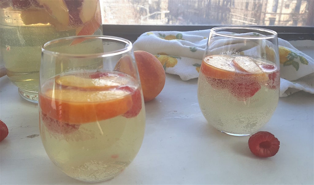 Peach sangria in a glad pitcher with fresh peaches and raspberries and a wooden spoon, next two more peach sangria and fruit in two glasses surrounded by fresh peaches and raspberries and a lemon dish towel on a white background. Front Shot with lace curtains. Sugar with Spice Blog.