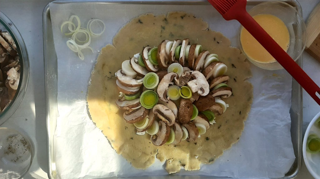 Overhead Mushroom Leek Galette raw all butter seasoned crust with mushrooms, leeks, bacon, and goat cheese assembled, ready for folding on an oven pan. Egg wash, mushrooms, and leeks in bowls scattered around.