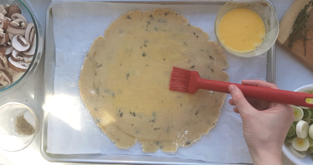 Overhead of raw all-butter seasoned crust on an oven sheet with an egg wash being applied with a red pastry brush. Leeks, mushrooms, thyme, salt, pepper, goat cheese, and egg wash surround it.