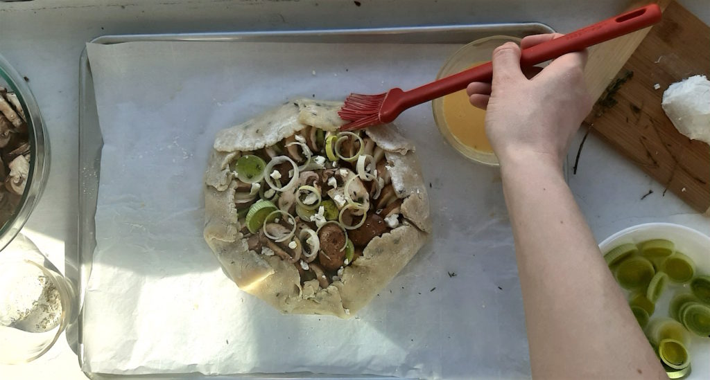 Raw mushroom and leek tart, stuffed and folded, on an oven sheet with a woman's hand applying eggwash over the crust with a red pastry brush