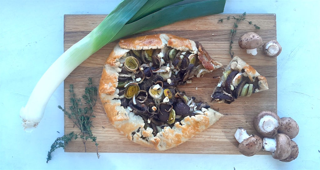 Overhead shot of Mushroom Leek Galette with goat cheese and thyme wrapped in all butter crust on a wooden cutting board. Surrounded by leeks, thyme and mushrooms. One slice cut out.