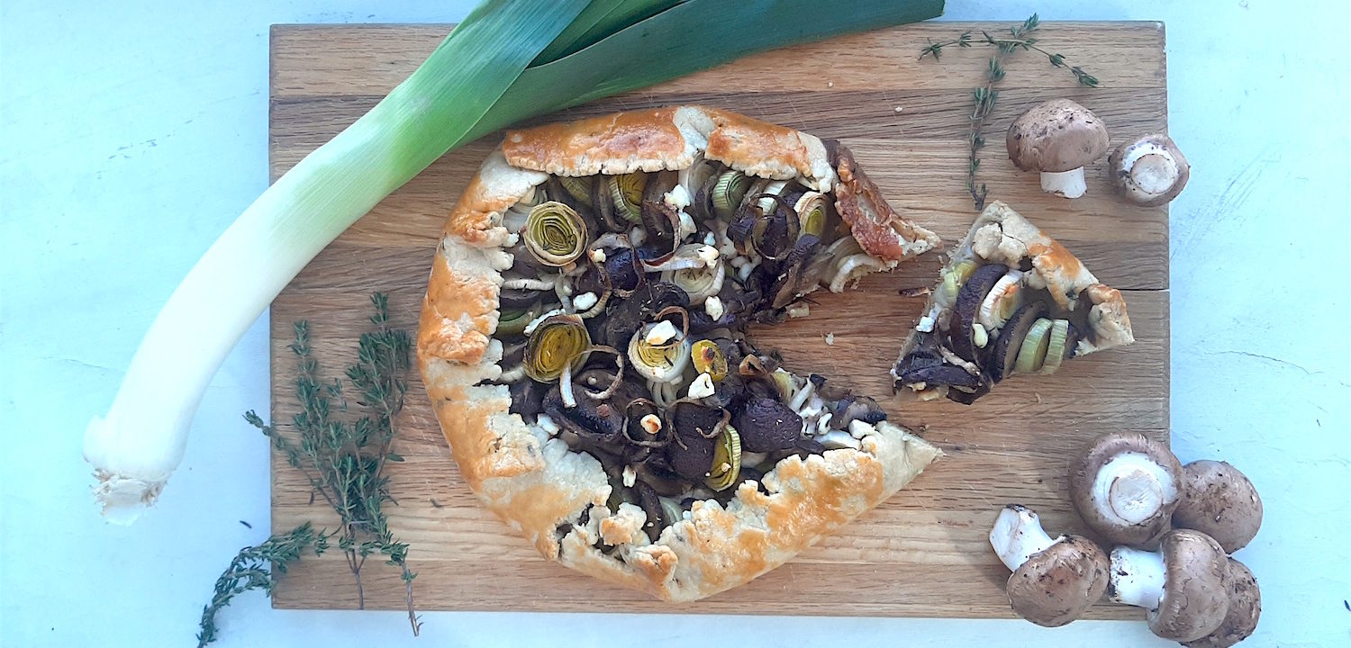 Overhead shot of Mushroom Leek Galette with goat cheese and thyme wrapped in all butter crust on a wooden cutting board. Surrounded by leeks, thyme and mushrooms. One slice cut out.