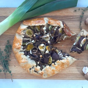 Overhead shot of Mushroom Leek Galette with goat cheese and thyme wrapped in all butter crust on a wooden cutting board. Surrounded by leeks, thyme and mushrooms. One slice cut out.