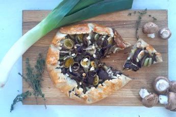 Overhead shot of Mushroom Leek Galette with goat cheese and thyme wrapped in all butter crust on a wooden cutting board. Surrounded by leeks, thyme and mushrooms. One slice cut out.