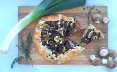 Overhead shot of Mushroom Leek Galette with goat cheese and thyme wrapped in all butter crust on a wooden cutting board. Surrounded by leeks, thyme and mushrooms. One slice cut out.