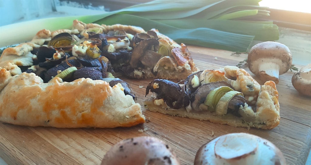 Side Shot Mushroom Leek Galette with goat cheese and thyme wrapped in all butter crust on a wooden cutting board. Surrounded by leeks, thyme and mushrooms.