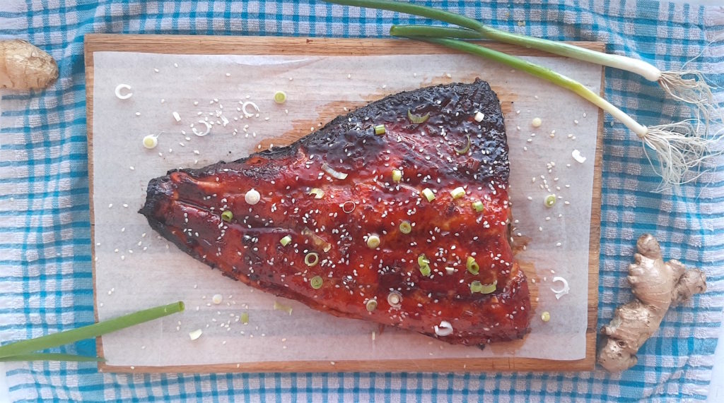 Honey and Wasabi Glazed Salmon on a wooden cutting board and blue checked dish towel surrounded by green scallions and fresh ginger and sesame seeds. Sugar with Spice Blog.