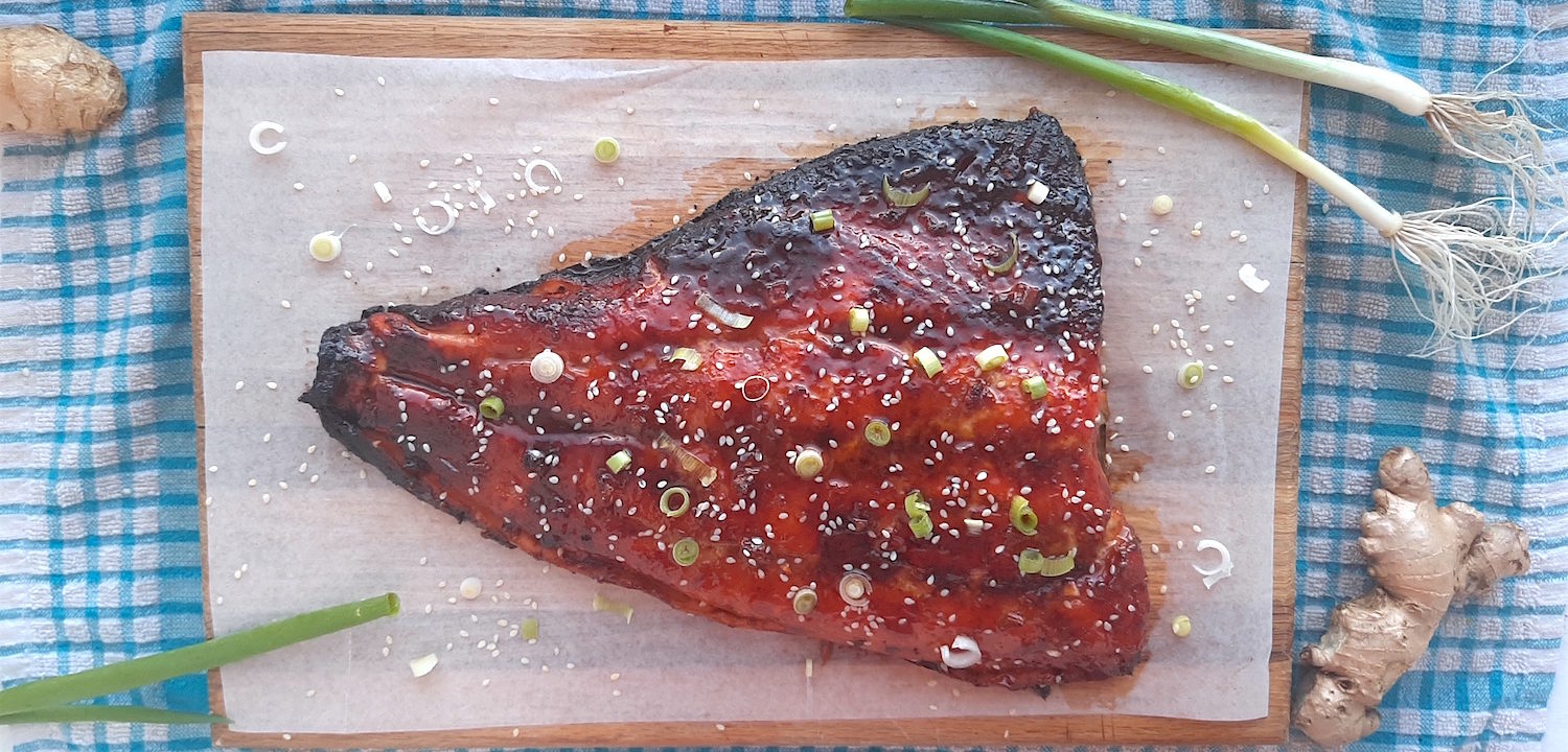 Honey and Wasabi Glazed Salmon on a wooden cutting board and blue checked dish towel surrounded by green scallions and fresh ginger and sesame seeds