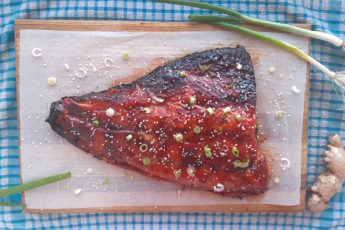 Honey and Wasabi Glazed Salmon on a wooden cutting board and blue checked dish towel surrounded by green scallions and fresh ginger and sesame seeds