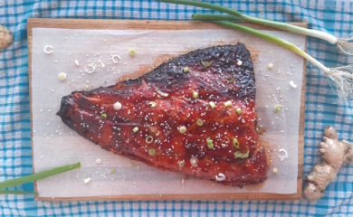 Honey and Wasabi Glazed Salmon on a wooden cutting board and blue checked dish towel surrounded by green scallions and fresh ginger and sesame seeds