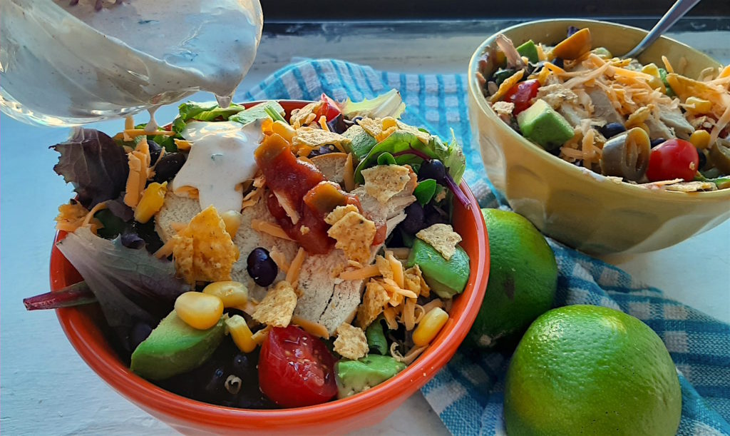 Creamy cilantro ranch dressing being poured from a glass cup over taco salad in an orange bowl. Taco salad in a yellow bowl in the background with blue checked dish towel and fresh limes on a white windowsill.