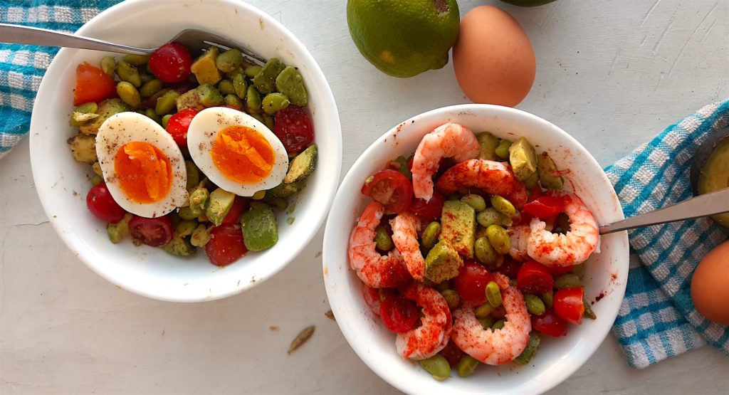 Protein Salad with edamame, sliced cherry tomatoes, avocado, eggs, and shrimp in two white bowls with silver forks on a white window sill with blue checked towels. Sugar with Spice Blog.