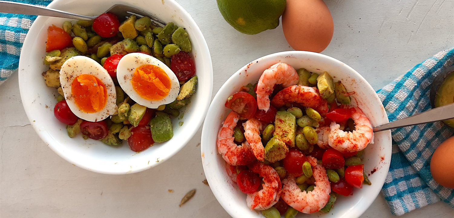 Protein Salad with edamame, sliced cherry tomatoes, avocado, eggs, and shrimp in two white bowls with silver forks on a white window sill with blue checked towels.