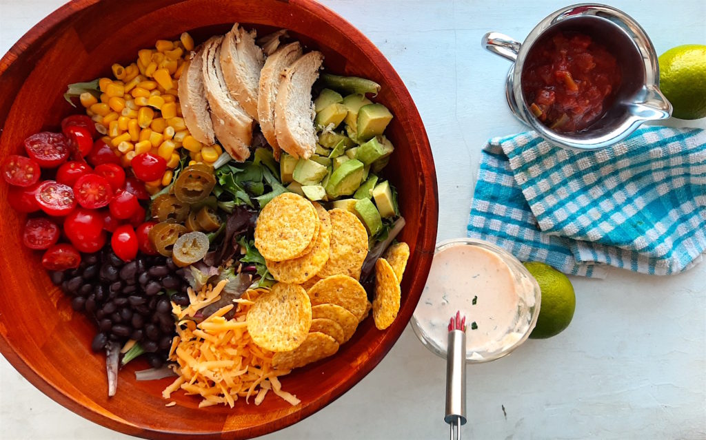 Sante Fe Taco Salad ingredients in a wooden salad bowl with tomatoes, black beans, cheddar cheese, tortilla chips, avocado, grilled chicken, corn, and greens. Creamy cilantro ranch in a glass dish and salsa in a metal dish with fresh green limes and blue checked dish towel on a white background.