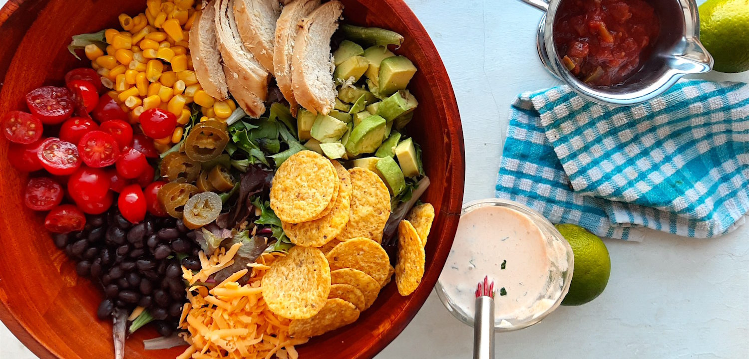 Sante Fe Taco Salad ingredients in a wooden salad bowl with tomatoes, black beans, cheddar cheese, tortilla chips, avocado, grilled chicken, corn, and greens. Creamy cilantro ranch in a glass dish and salsa in a metal dish with fresh green limes and blue checked dish towel on a white background.