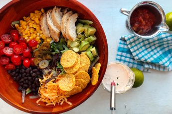 Sante Fe Taco Salad ingredients in a wooden salad bowl with tomatoes, black beans, cheddar cheese, tortilla chips, avocado, grilled chicken, corn, and greens. Creamy cilantro ranch in a glass dish and salsa in a metal dish with fresh green limes and blue checked dish towel on a white background.