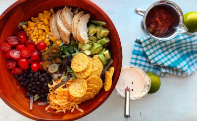 Sante Fe Taco Salad ingredients in a wooden salad bowl with tomatoes, black beans, cheddar cheese, tortilla chips, avocado, grilled chicken, corn, and greens. Creamy cilantro ranch in a glass dish and salsa in a metal dish with fresh green limes and blue checked dish towel on a white background.