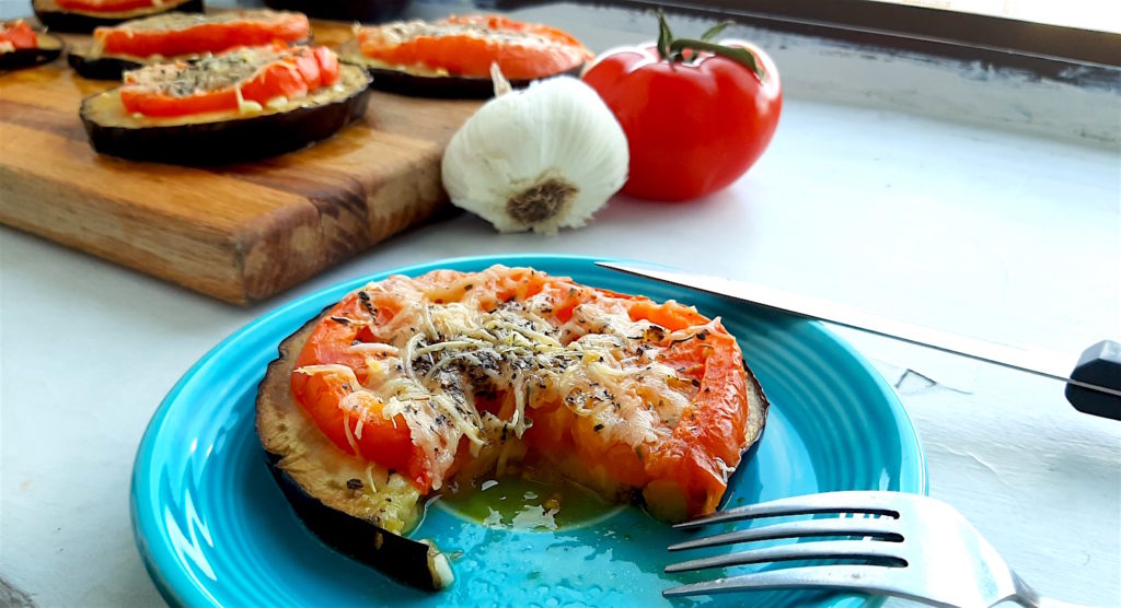 Low-carb eggplant and tomato pizza on a blue plate with one slice cut out, fork and knife nearby. More eggplant and tomato pizzas in the background on a wooden cutting board surrounded by tomatoes and garlic. Sugar with Spice Blog.