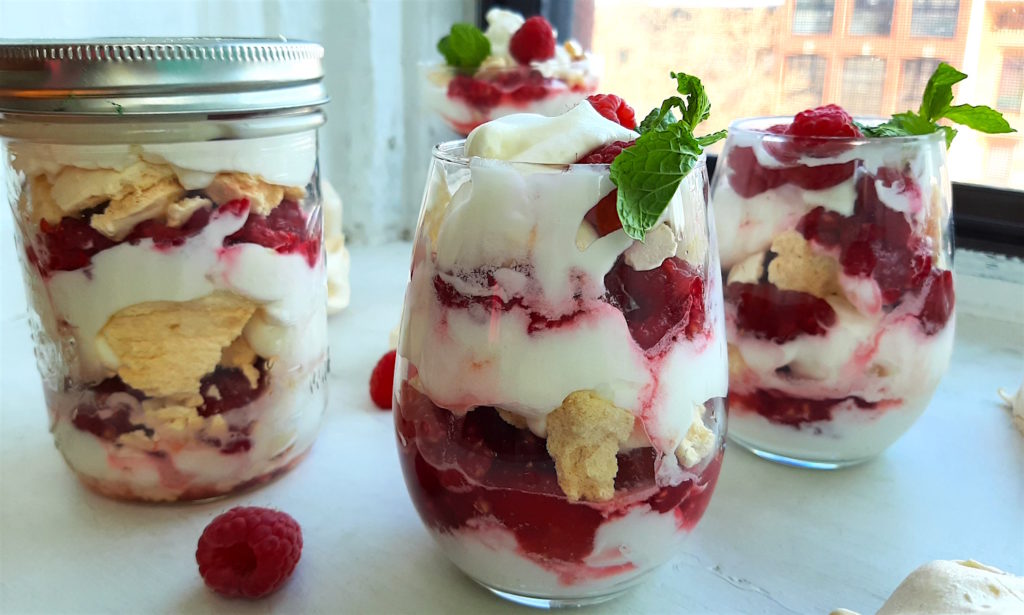 Boozy Raspberry Eaton Mess in two stemless wine glasses with mint leaf garnish. Mason Jar of Eton Mess in the background with meringue cookies. All on a white window sill. Sugar with Spice Blog.