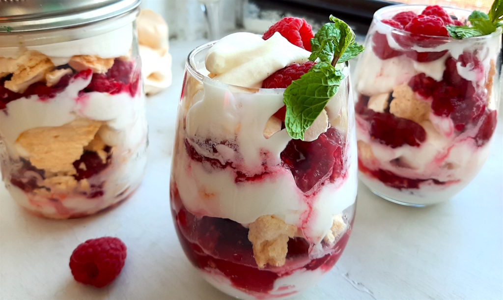 Boozy Raspberry Eaton Mess in two stemless wine glasses with mint leaf garnish. Mason Jar of Eton Mess in the background with meringue cookies. All on a white window sill. Sugar with Spice Blog.