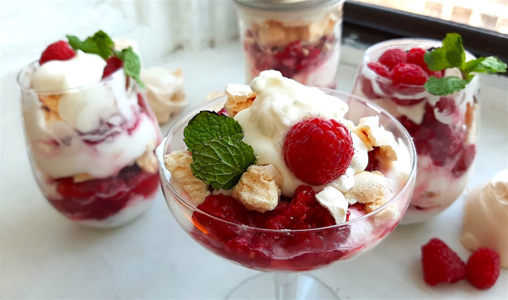 Boozy Raspberry Eaton Mess in a coupe  with mint leaf garnish. Two stemless wine glasses of Eton Mess and a closed mason jar of Eton Mess in the background with meringue cookies and raspberries. All on a white window sill. Sugar with Spice Blog.