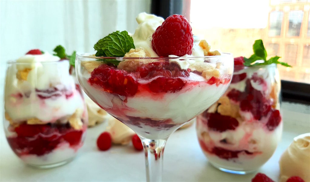 Boozy Raspberry Eaton Mess in a coupe with mint leaf garnish. Two stemless wine glasses of Eton Mess and a closed mason jar of Eton Mess in the background with meringue cookies and raspberries. All on a white window sill. Sugar with Spice Blog.