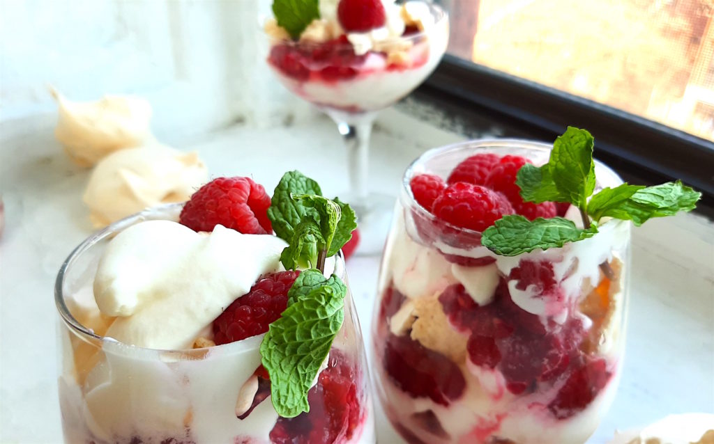 Boozy Raspberry Eaton Mess in two stemless wine glasses with mint leaf garnish. Coupe of Eton Mess in the background with meringue cookies. All on a white window sill. Sugar with Spice Blog.