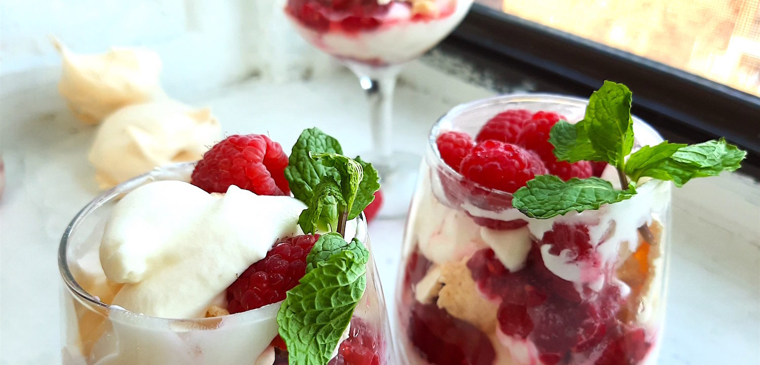 Boozy Raspberry Eaton Mess in two stemless wine glasses with mint leaf garnish. Coupe of Eton Mess in the background with meringue cookies. All on a white window sill. Sugar with Spice Blog.