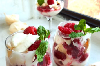 Boozy Raspberry Eaton Mess in two stemless wine glasses with mint leaf garnish. Coupe of Eton Mess in the background with meringue cookies. All on a white window sill. Sugar with Spice Blog.