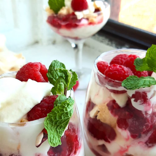Boozy Raspberry Eaton Mess in two stemless wine glasses with mint leaf garnish. Coupe of Eton Mess in the background with meringue cookies. All on a white window sill. Sugar with Spice Blog.