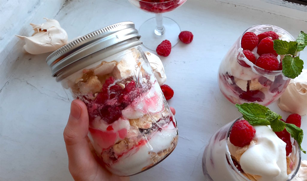 Mason Jar of Boozy Raspberry Eton Mess held in a woman's hand over a white window sill. Two stemless wine glasses and a coupe of eton mess with mint garnish and loose raspberries and meringue cookies in the background. Sugar with Spice Blog.