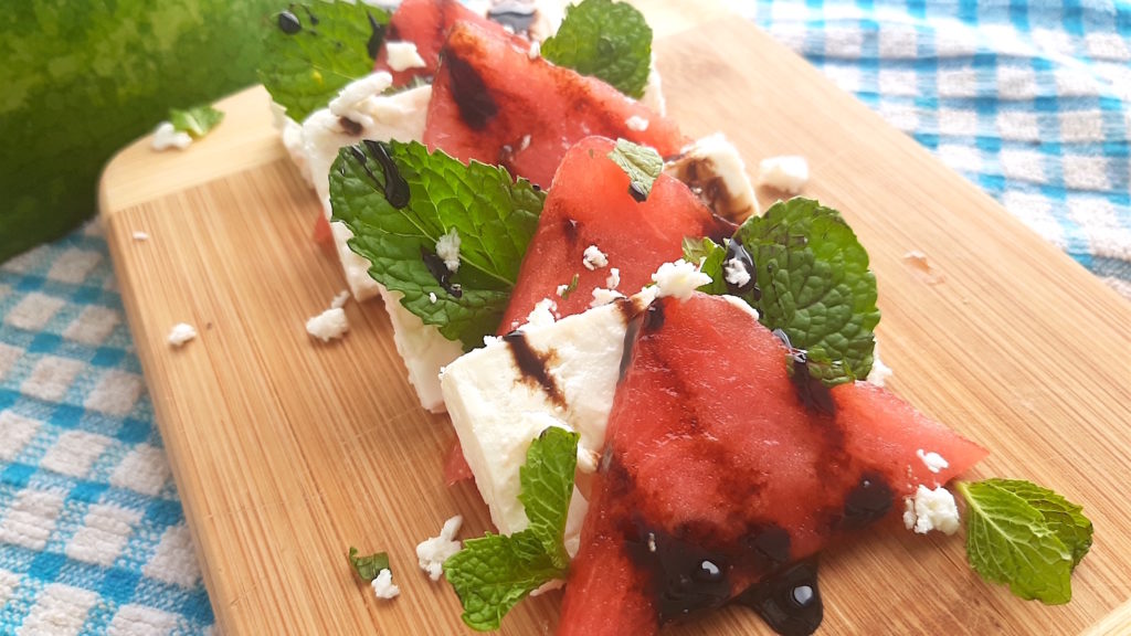 Summer watermelon salad fancy presentation with triangles of watermelon, feta, mint leaves and drizzled balsamic glaze on a small wooden cutting board with watermelon rind, and blue checked cloth in the background. Sugar with Spice Blog.