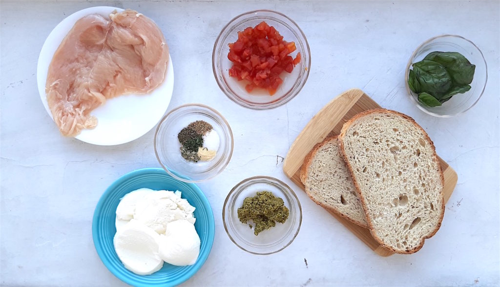 Chicken Caprese Panini Ingredients on a white window sill. Fresh chicken breast, seasoning, sliced fresh mozzarella, crushed red tomatoes, pesto, and fresh basil with two slices peasant bread. Sugar with Spice Blog.