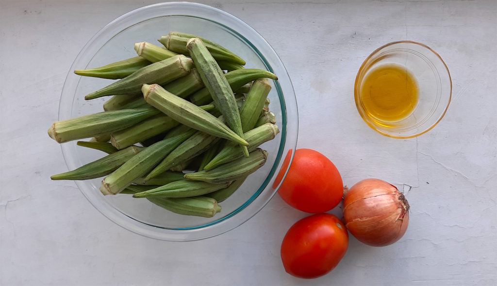 Southern Sautéed okra ingredients. Whole Okra in a pyrex bowl, whole plum tomatoes, whole onion, and olive oil in pyrex on a white background.