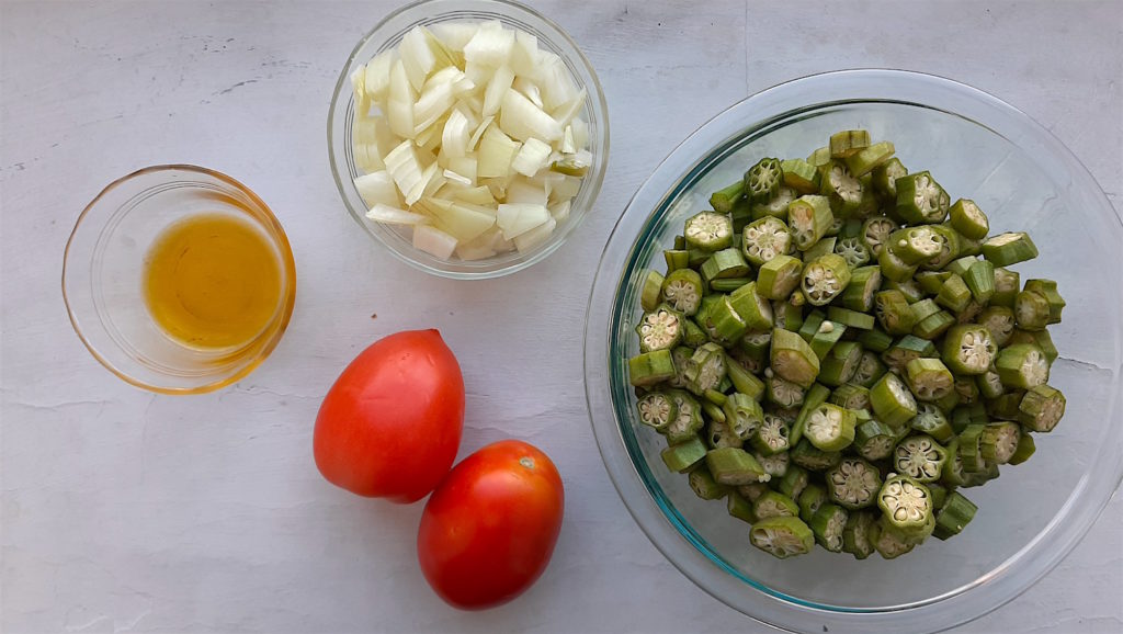 Southern Sautéed okra ingredients. Sliced Okra in a pyrex bowl, whole plum tomatoes, sliced white onion in pyrex, and olive oil in pyrex on a white background.