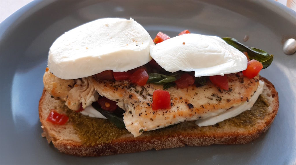 Chicken Caprese Panini almost final step. Bottom layer of bread with pesto, crushed tomatoes, fresh basil and mozzarella layered on seasoned chicken breast in a non stick frying pan. Sugar with Spice Blog.