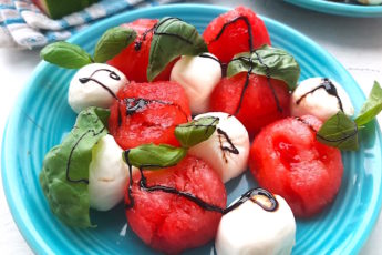 Summer Watermelon Salad close up of watermelon balls, mozzarella balls, basil leaves, balsamic reduction on a blue plate. Sugar with Spice Blog.