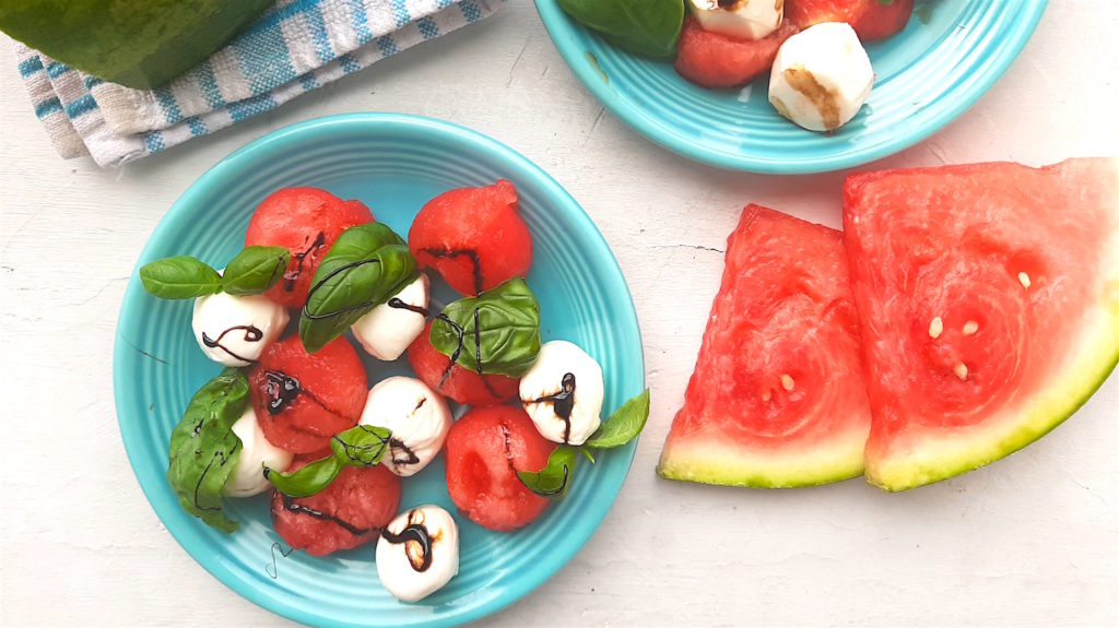 Summery Watermelon Salad second version with mozzarella balls, watermelon balls, basil leaves, and balsamic reduction. On two blue plates, over head shot with watermelon slices and blue checked cloth in the background on a white window sill. Sugar with Spice Blog.