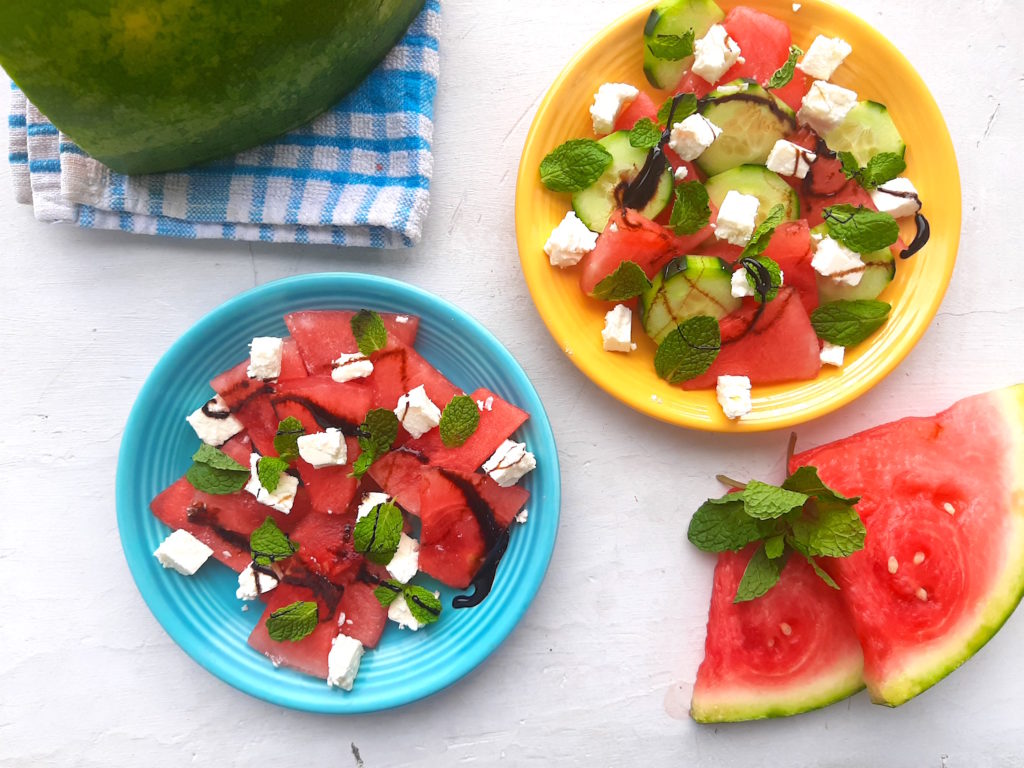 Summery Watermelon Salad with mint, feta, balsamic reduction, and cucumber on blue and yellow plates with sliced watermelon and blue checked cloth in the background. Overhead shot on a white window sill. Sugar with Spice Blog.