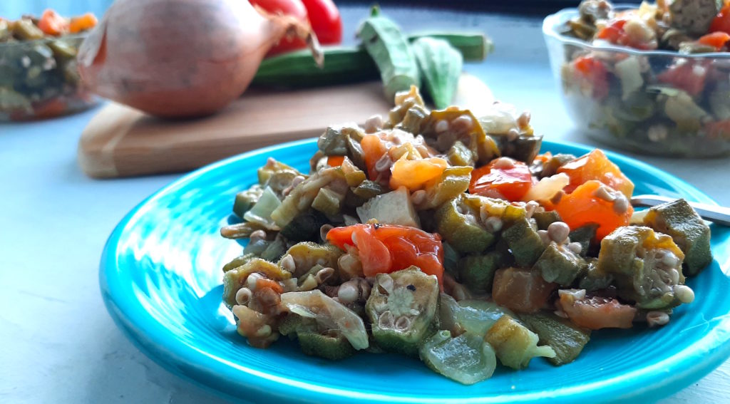 Southern Sautéed okra with onions, garlic, and tomatoes on a blue plate, front shot. Glass pyrex bowls with more okra, tomatoes, garlic, and onions in the background. Also a wooden cutting board with a whole onion, whole okra, and tomatoes in the background. Sugar with Spice Blog.